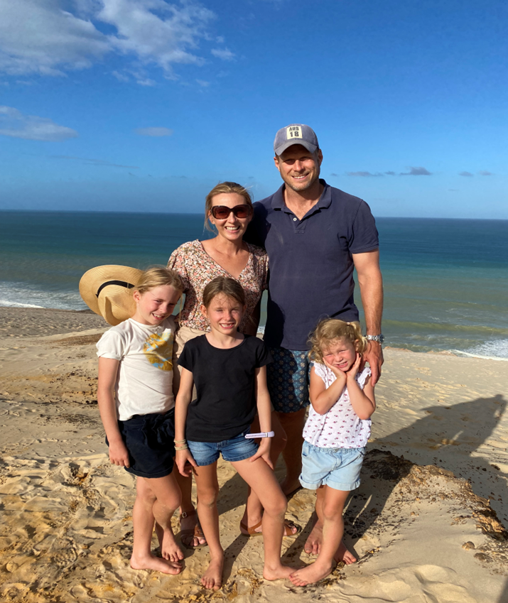 A family stands together on the beach, smiling for a photo with the ocean waves in the background.