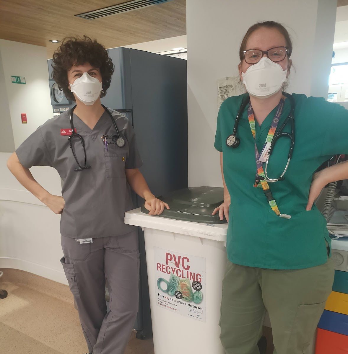 Two women in scrubs stand beside a trash can, engaged in conversation in a healthcare setting.