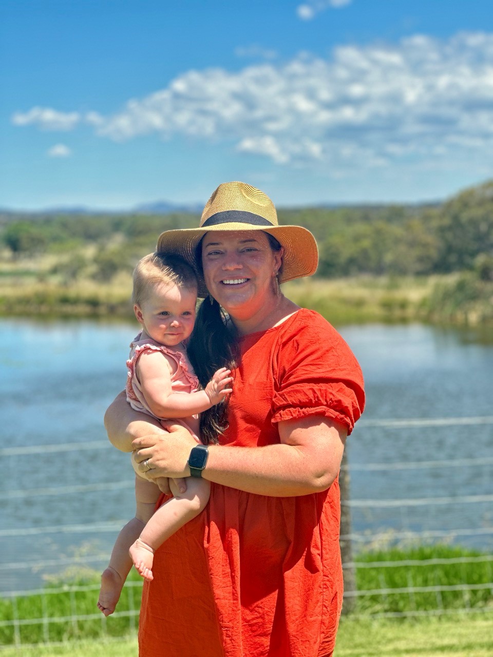 A woman in a red dress cradles a baby in her arms while standing in a lush green field under a clear blue sky.