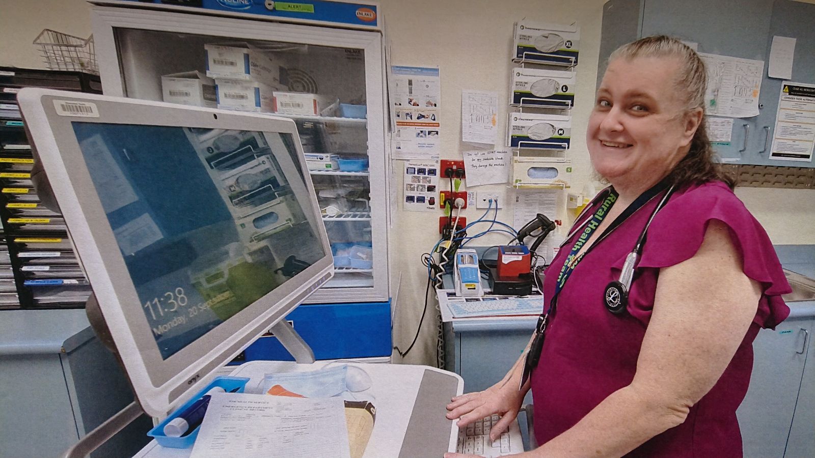 A woman in a hospital room, engaged with a computer, with medical supplies and a bed visible in the background.