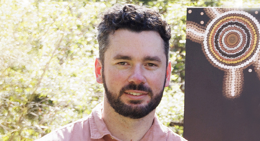 Jacob PAGE with a beard standing next to a First Nation artwork.