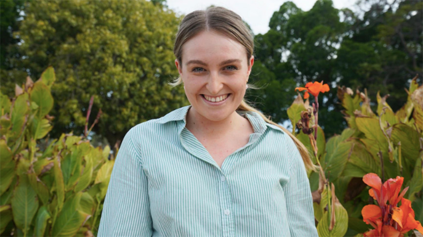 A woman in a green shirt stands gracefully in front of a vibrant display of blooming flowers.